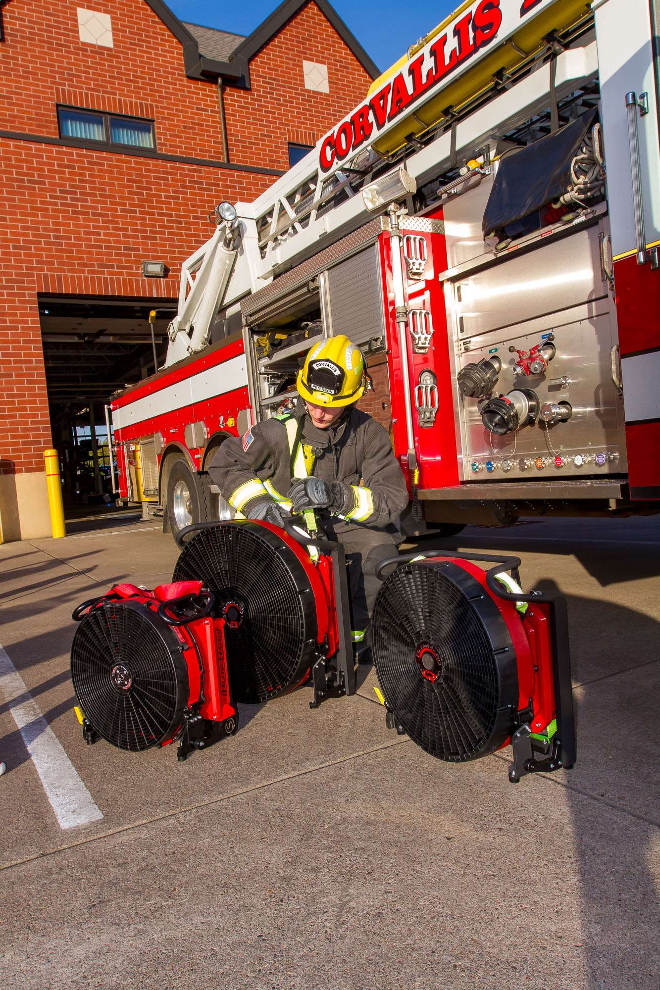 A firefighter in full protective gear kneeling behind BlowHard's Squirt, Quickee, and Commando PPV fan models, adjusting the Commando model.