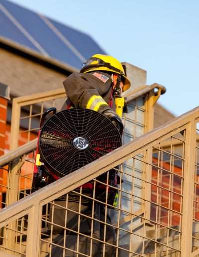 A firefighter walking up a staircase with the Squirt BlowHard fan model strapped over his right shoulder.